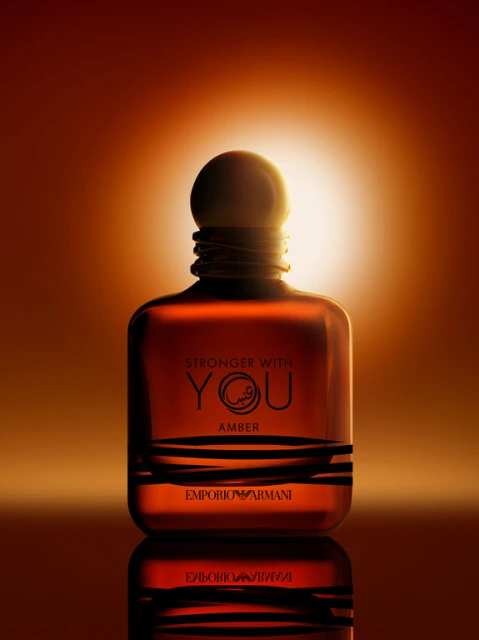 Amber-toned glass perfume bottle with black accents, lit dramatically against a gradient background in shades of orange and yellow.