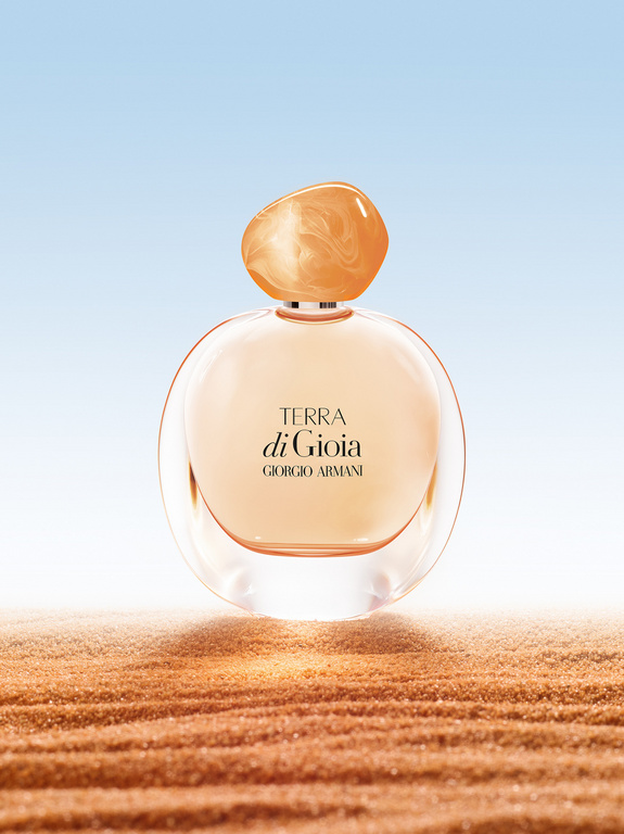 Close-up product shot of Giorgio Armani 'Terra di Gioia' perfume bottle with a round shape and orange lid on sand against blue sky.