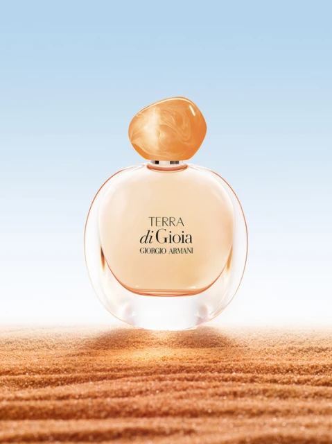 Close-up product shot of Giorgio Armani 'Terra di Gioia' perfume bottle with a round shape and orange lid on sand against blue sky.