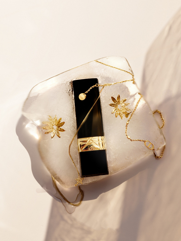 Elegant beauty still life featuring a black lipstick tube with gold floral accents and chain on a clear textured surface.