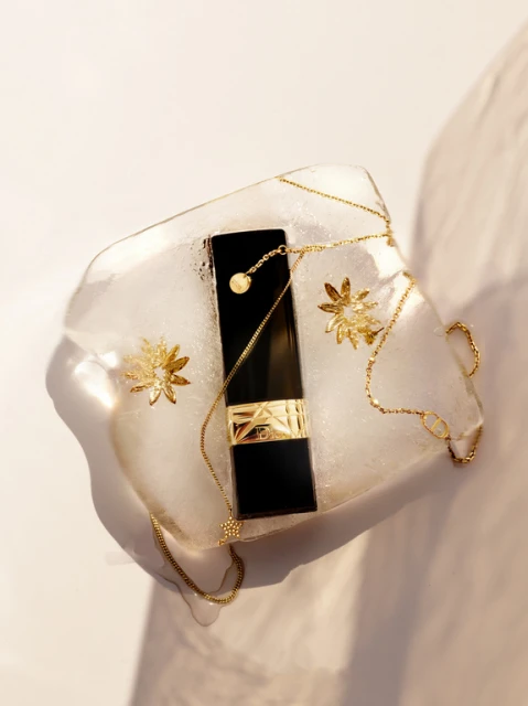 Elegant beauty still life featuring a black lipstick tube with gold floral accents and chain on a clear textured surface.