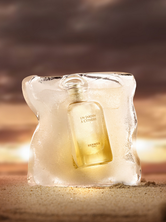 Hermès Un Jardin à Cythère perfume bottle encased in ice sits on a sandy surface with a blurred brown and gold sunset background.