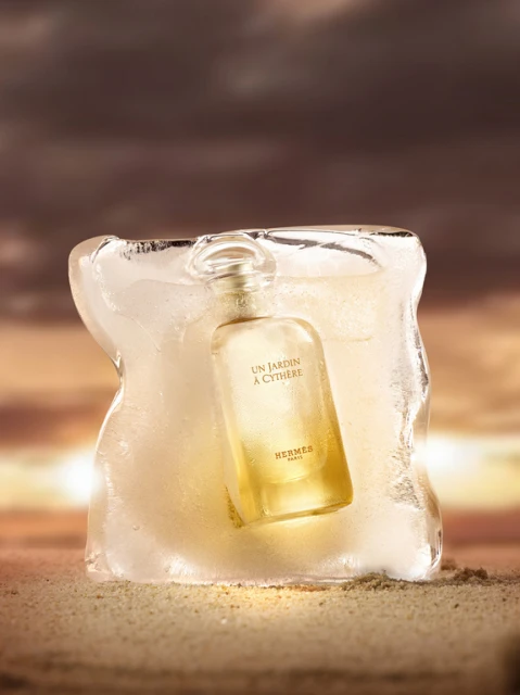 Hermès Un Jardin à Cythère perfume bottle encased in ice sits on a sandy surface with a blurred brown and gold sunset background.