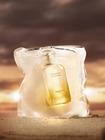 Hermès Un Jardin à Cythère perfume bottle encased in ice sits on a sandy surface with a blurred brown and gold sunset background.