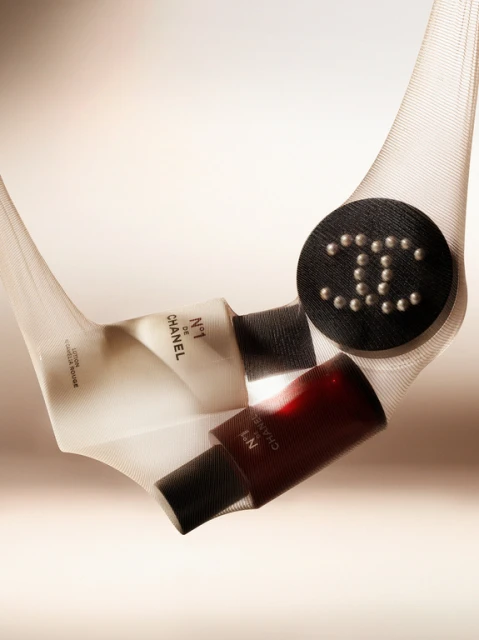 Still life featuring Chanel No. 1 de Chanel skincare and makeup products in a white mesh sling, against a soft peach background.