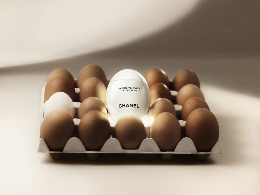 Cosmetic product placed within an egg carton, surrounded by brown and one white egg against a pale, illuminated backdrop.