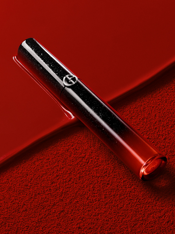 Close-up of a gradient red and black Giorgio Armani lip gloss tube, positioned diagonally on a textured red background for beauty.