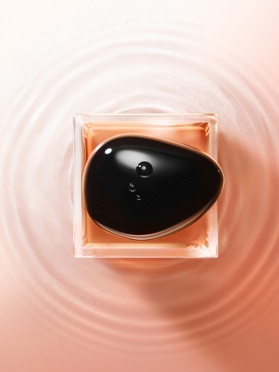 Close-up overhead shot of clear, square perfume bottle with brown liquid, topped by a glossy black pebble, against a peach background with water ripples.