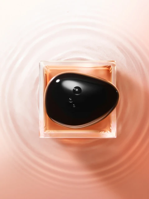 Close-up overhead shot of clear, square perfume bottle with brown liquid, topped by a glossy black pebble, against a peach background with water ripples.