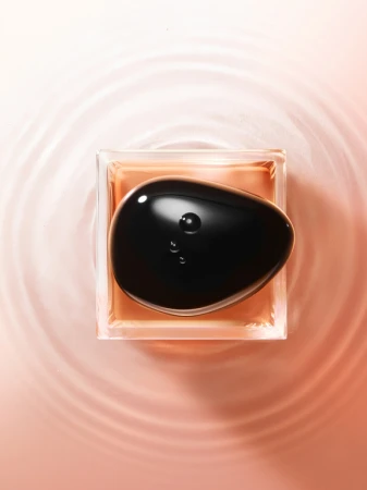 Top-down view of a dark polished smooth stone resting inside a square glass bottle filled with amber liquid on rippled pink water.