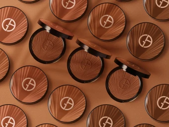 A flat lay arrangement of multiple round bronze makeup compacts with sunburst patterns on a warm brown background.