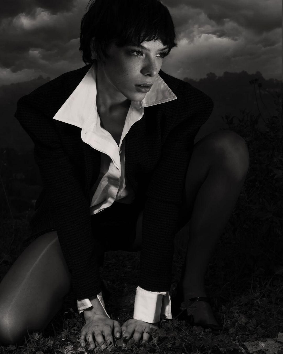 Monochrome portrait of a crouched adult female in a black blazer and white shirt against a stormy sky background, gazing away.