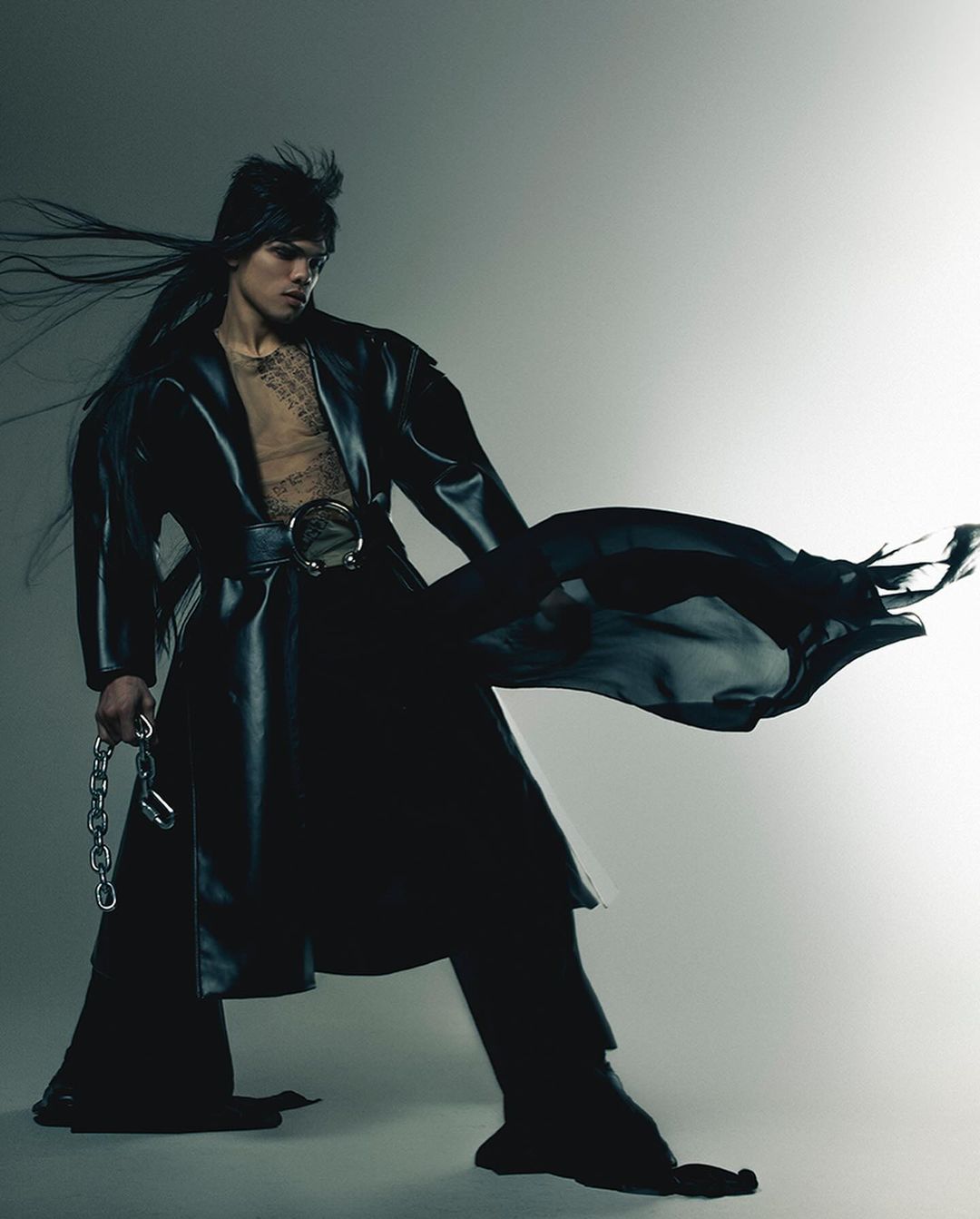 A person in dark, stylish clothing poses dramatically against a gradient gray backdrop, showcasing a bold and modern fashion aesthetic.