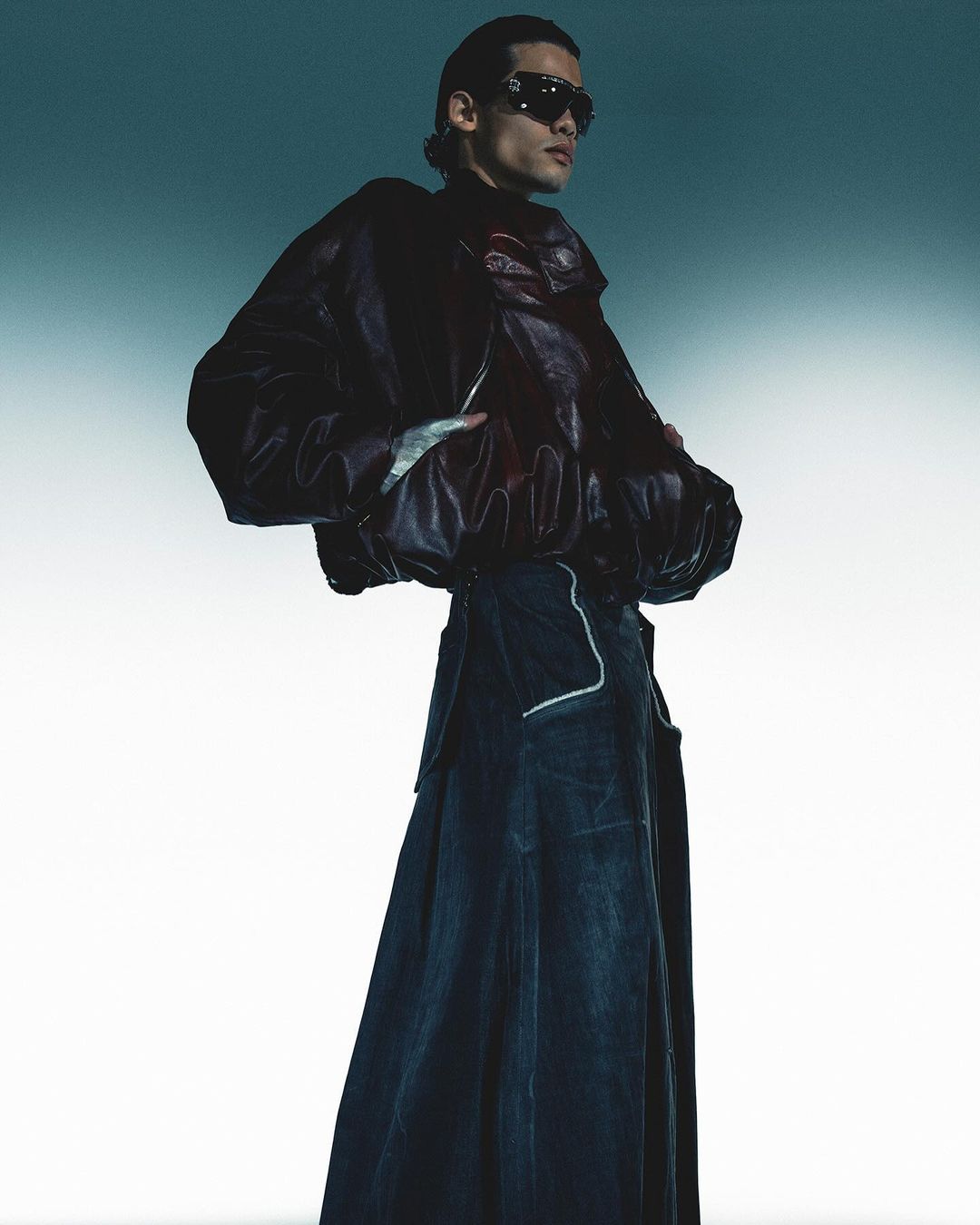 A lone adult poses in a studio, wearing a black leather jacket and long denim skirt, complemented by dark sunglasses and a light grey backdrop.