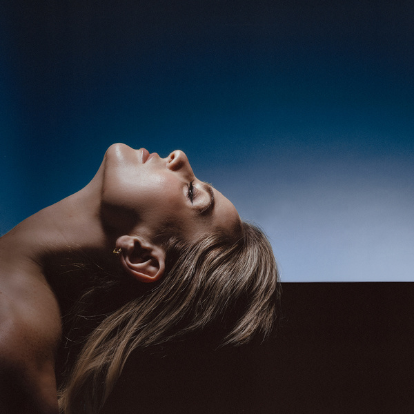 Portrait of a young adult woman with blonde hair tilted upward against a dark blue background in an indoor studio setting.