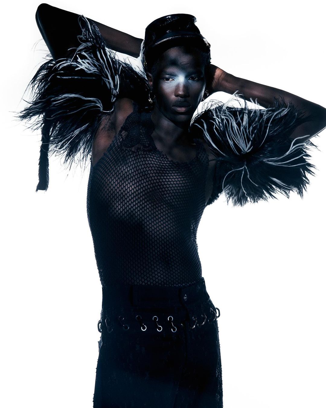 Fashionable woman with dark skin poses with hands on her head in a black net outfit with feathered epaulettes against a white backdrop.