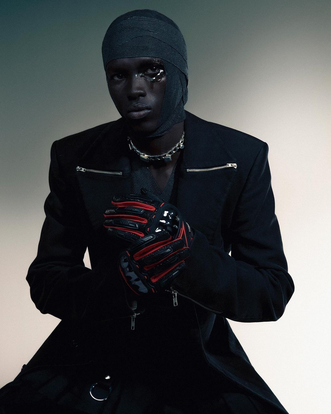 Fashion portrait of a black male model with head wrapped, wearing black coat, red/black gloves and silver chain, against a blurred background.