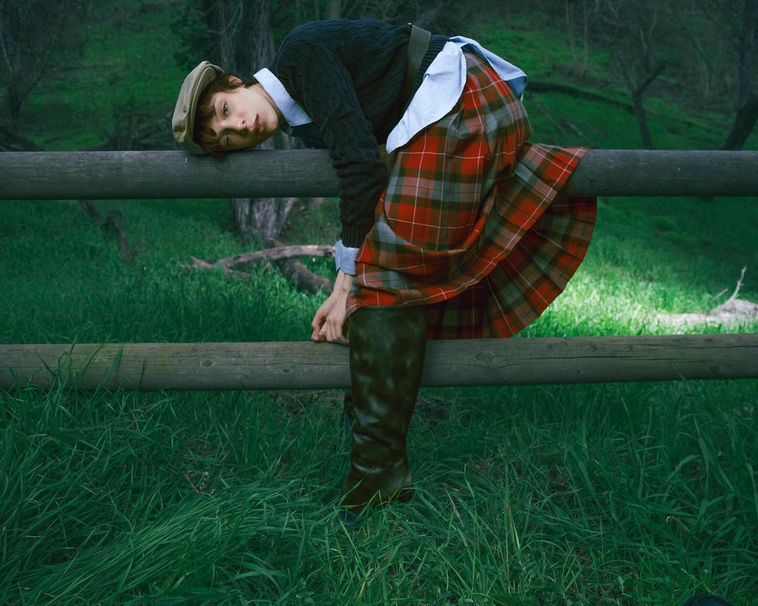 An adult with a hat rests on a wooden fence, wearing a black sweater, white collared shirt, and a red and green plaid skirt in a field.