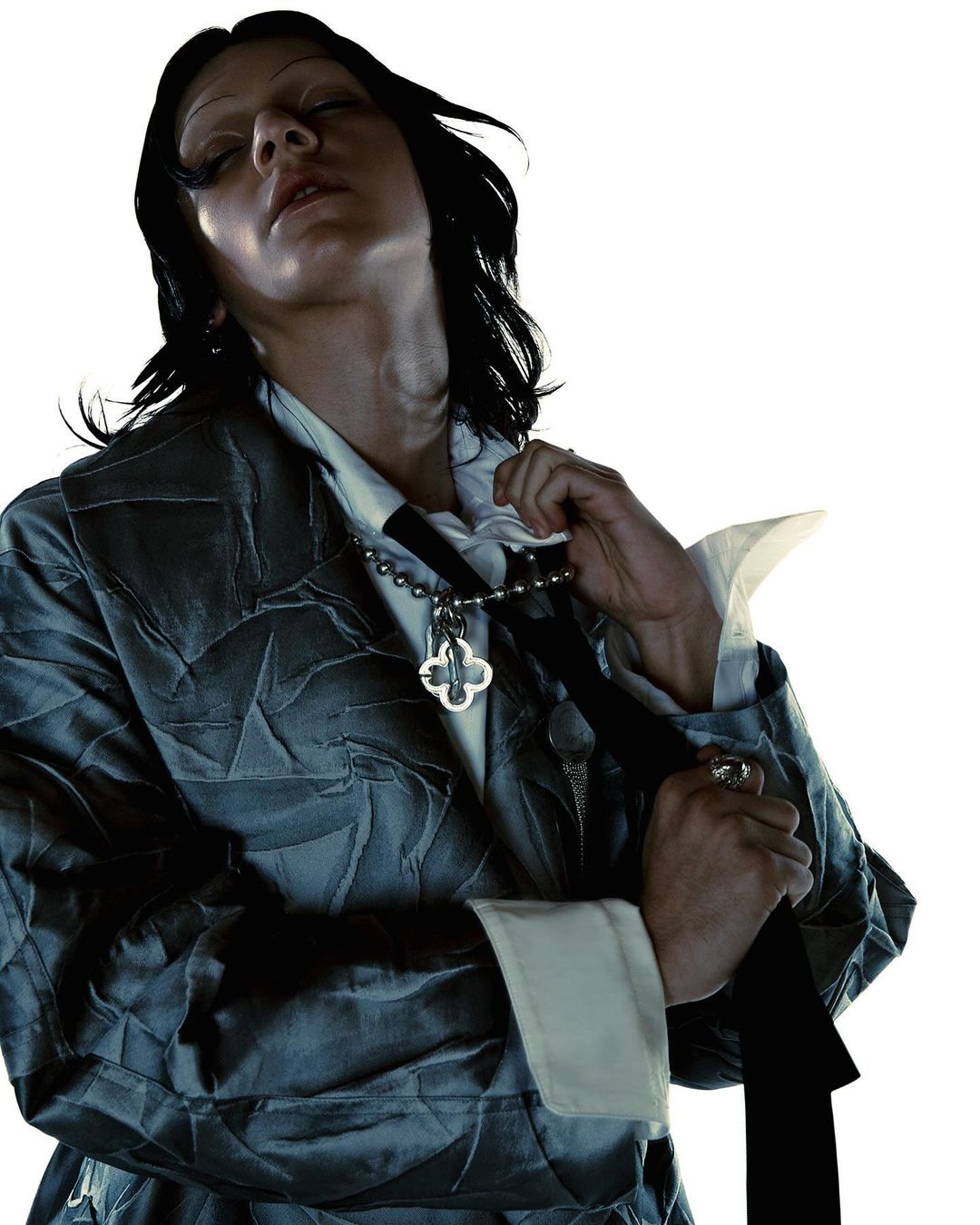 A person in a patterned jacket and tie with a quatrefoil pendant, eyes closed, poses in a brightly lit, high-key portrait.