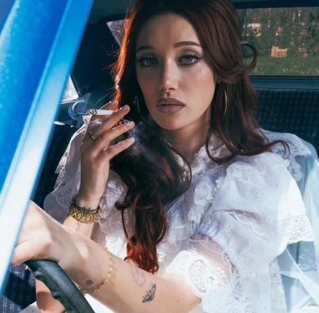 Woman in a car smoking a cigarette. She has long, auburn hair and is wearing a white lace dress with a blue interior car.