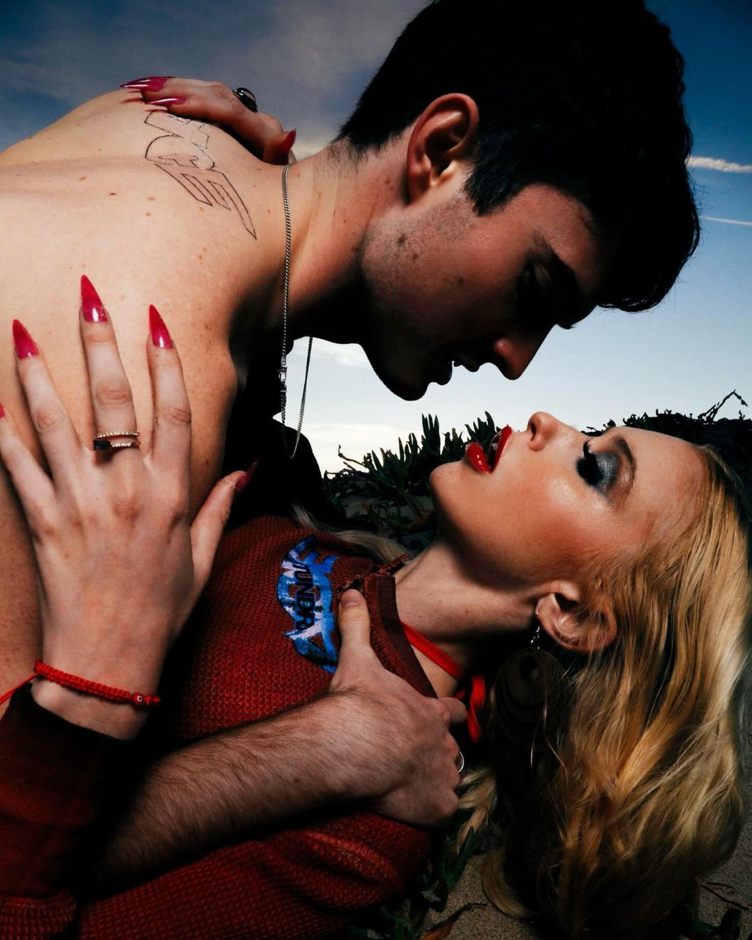Close up portrait of a shirtless male and a blond woman lying down. They are embraced in an intimate position, with a sky background.