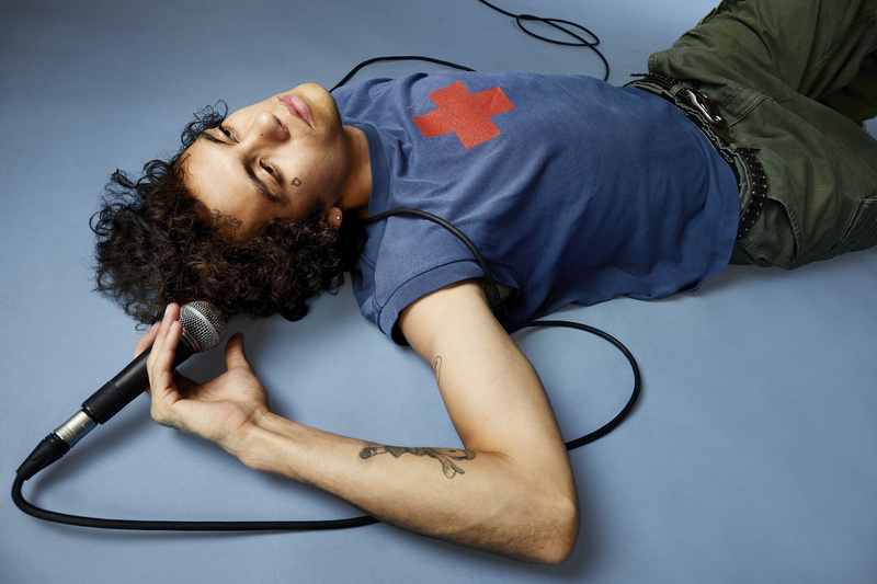Man with curly hair and a red cross t-shirt lies on a blue surface holding a microphone, surrounded by a black cable in a studio setting.