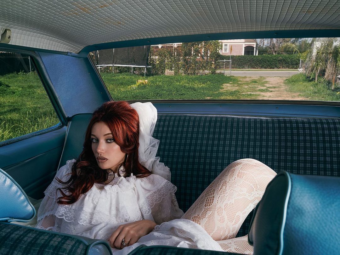 Portrait of a red-haired woman in a vintage car wearing a white lace dress, white stockings, and large hoop earrings, with a house in background.