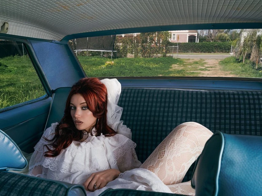 Portrait of a red-haired woman in a vintage car wearing a white lace dress, white stockings, and large hoop earrings, with a house in background.