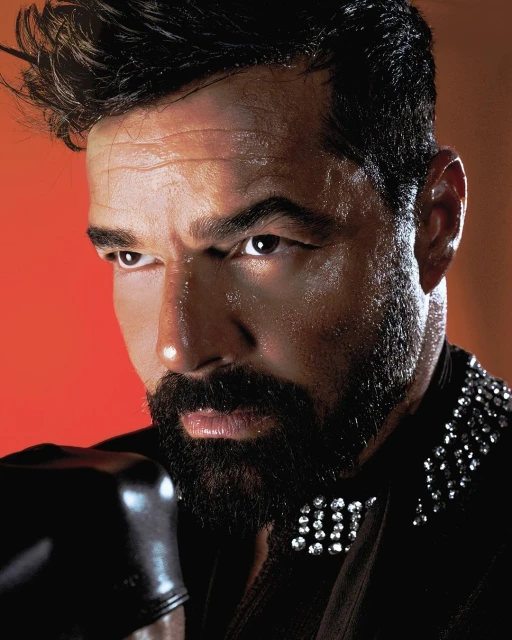 Close-up of an adult male with a beard wearing a black jeweled jacket and boxing gloves, set against an orange studio backdrop with hard lighting.