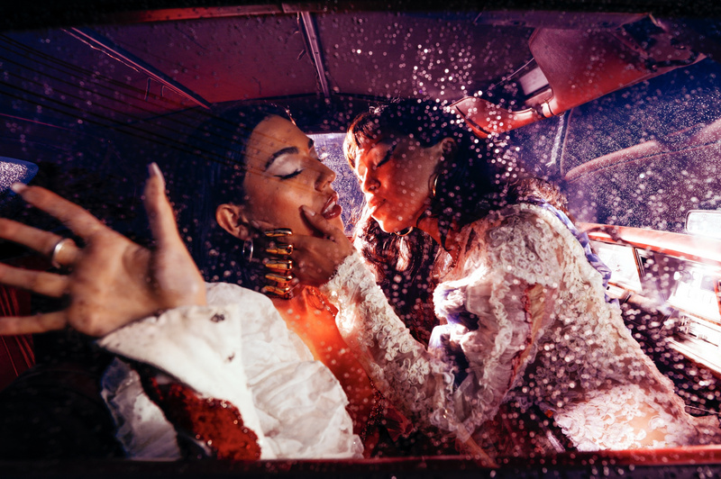 Two women in vintage dresses share a tender moment inside a vintage car, with rain or water droplets visible on the car windows.