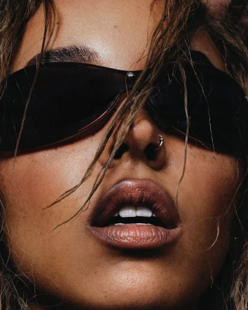 Close-up portrait of a person wearing dark wraparound sunglasses with wet hair strands framing their face and nose ring.