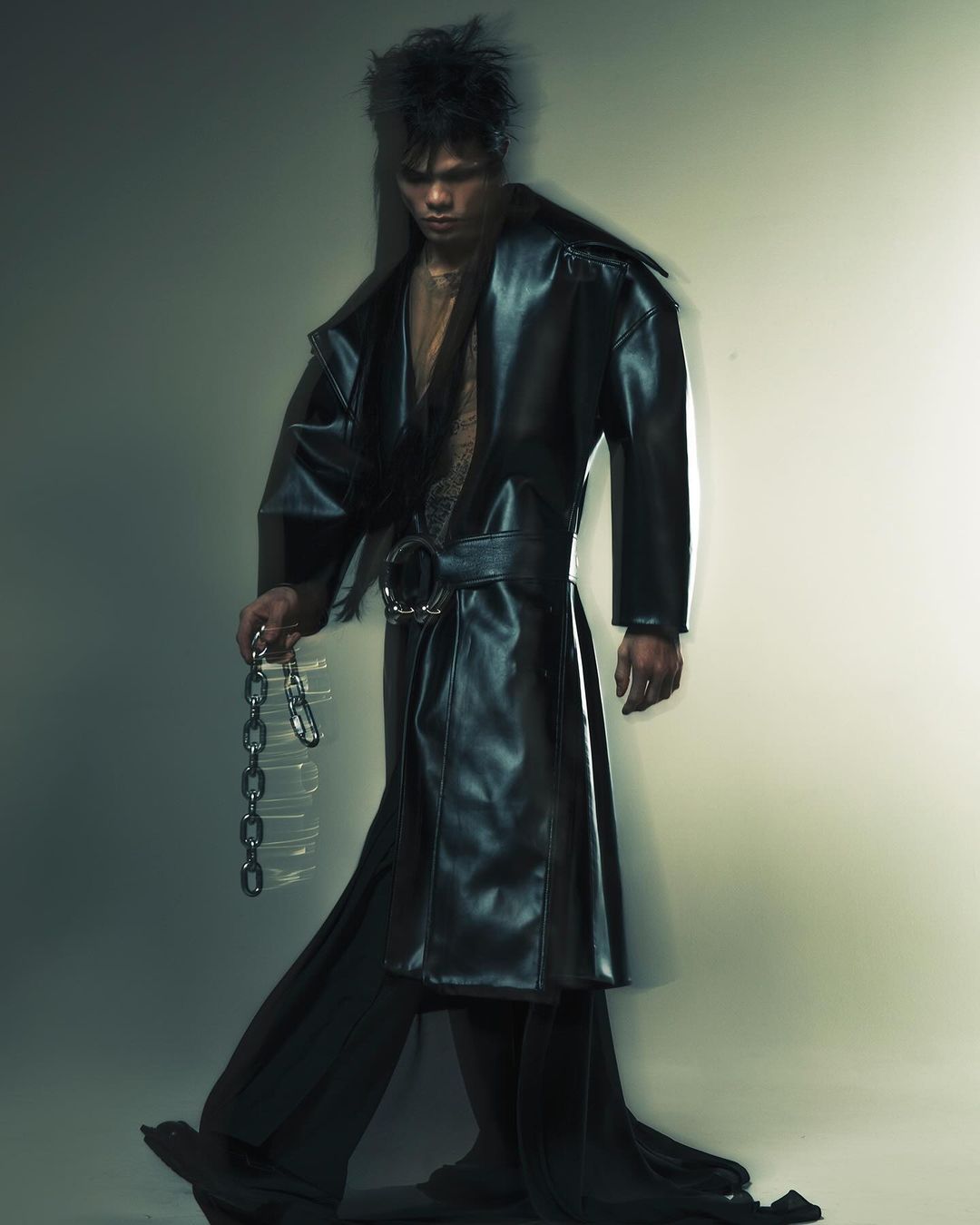 A person in a long black coat and wide-leg pants walks holding a metal chain against a graded light gray backdrop in a studio shot.