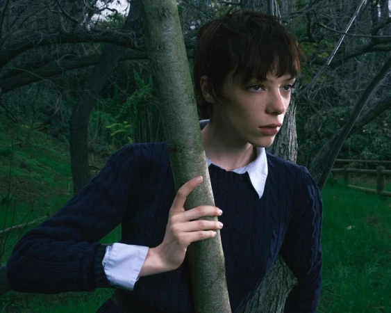 Short-haired woman in a dark blue sweater holding a tree trunk outdoors in a forest, photographed by Callum Walker Hutchinson.