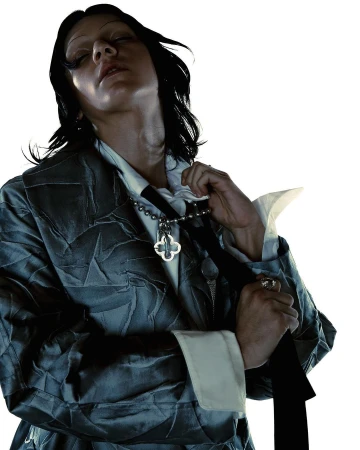 Model with dark hair and closed eyes wearing a crumpled grey coat, white shirt, and a clover necklace against a bright background.