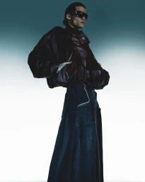 Model in dark sunglasses wearing a glossy dark leather jacket and denim skirt against a gradient background by Callum Walker Hutchinson