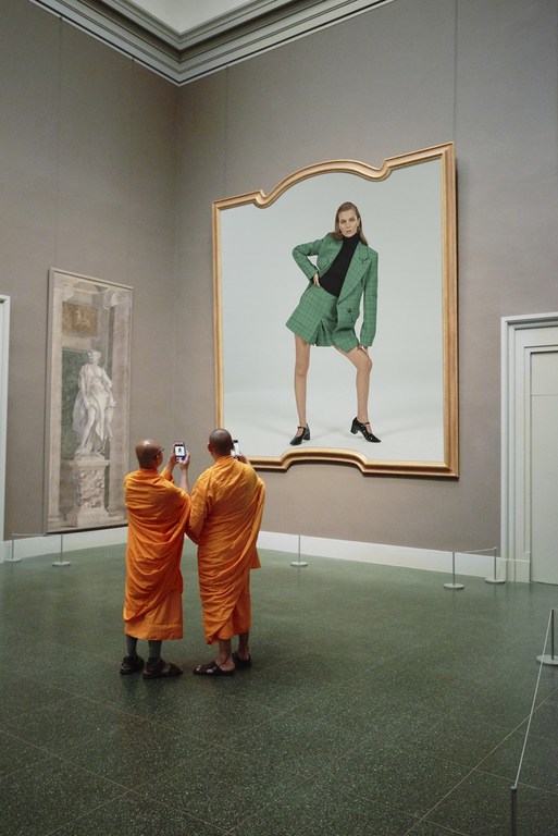 Two monks photograph a woman in a green suit inside a large gold frame inside an art museum. Visitors, art and fashion elements.