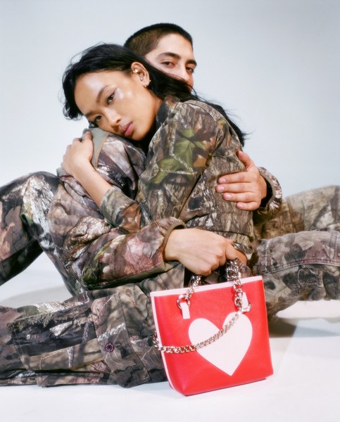 An Asian woman and a Latino man pose in matching camouflage outfits, highlighting a red and white heart-shaped handbag, set against a studio backdrop.