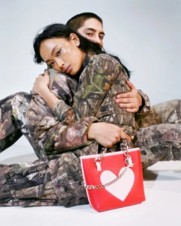 An Asian woman and a Latino man pose in matching camouflage outfits, highlighting a red and white heart-shaped handbag, set against a studio backdrop.