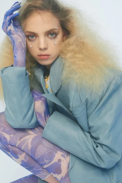 Woman in blue jacket and floral tights poses in photo studio with theatrical tears. Her blonde hair is wavy and slightly disheveled.