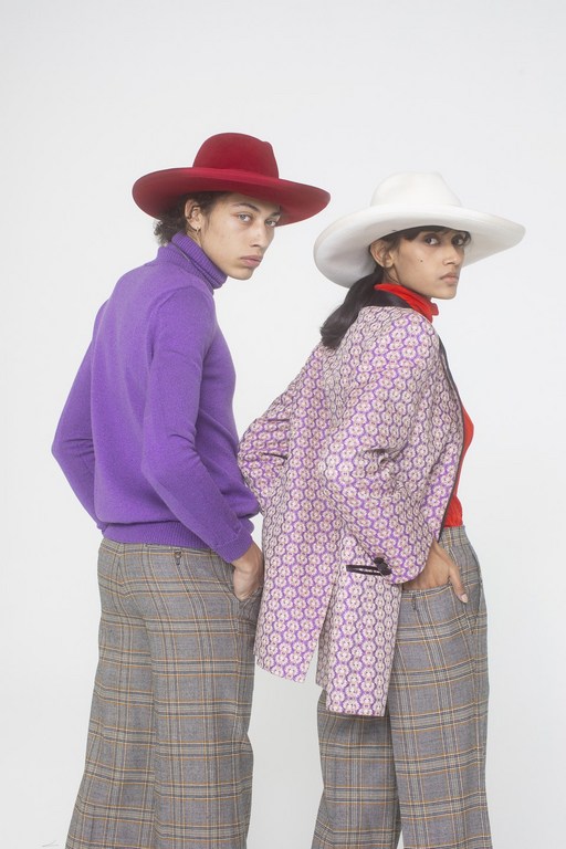 Two fashion models in wide-leg plaid pants and hats pose in a photo studio. One wears a purple sweater and red hat; the other a patterned blazer.
