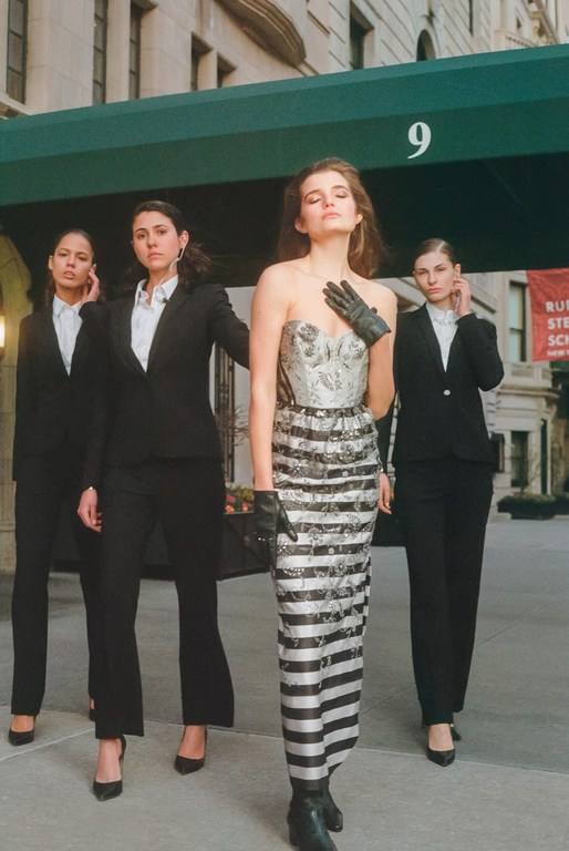 Fashion shot of a woman in a strapless dress with a striped skirt, accompanied by three women in black suits outdoors in daytime.