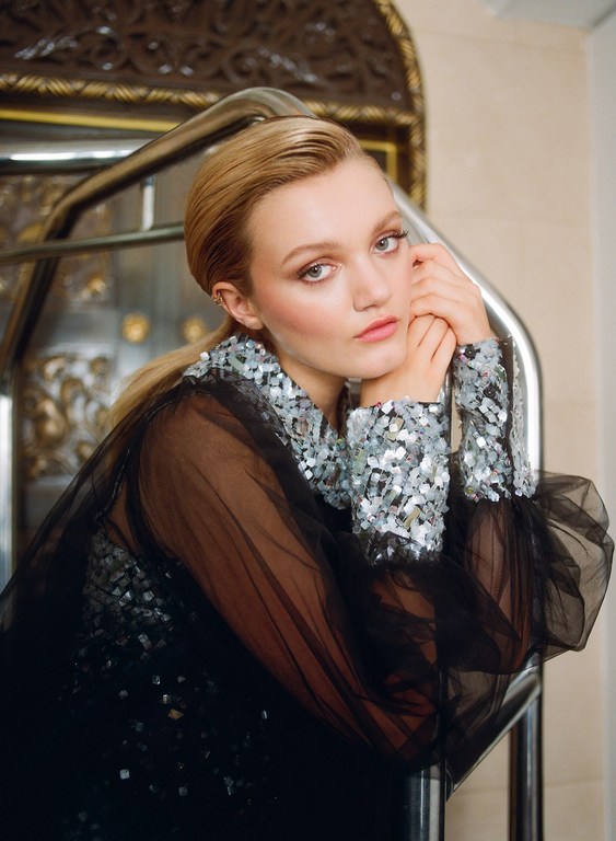 Blonde woman with sequin top and black tulle sleeves poses near a metal structure inside an ornately decorated interior location.