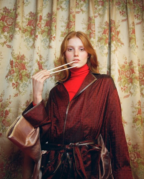 Young adult woman poses indoors with chopsticks in front of floral patterned curtains, wearing a red turtleneck and a burgundy robe.