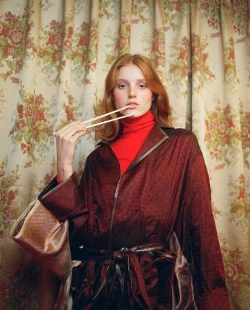 Young adult woman poses indoors with chopsticks in front of floral patterned curtains, wearing a red turtleneck and a burgundy robe.