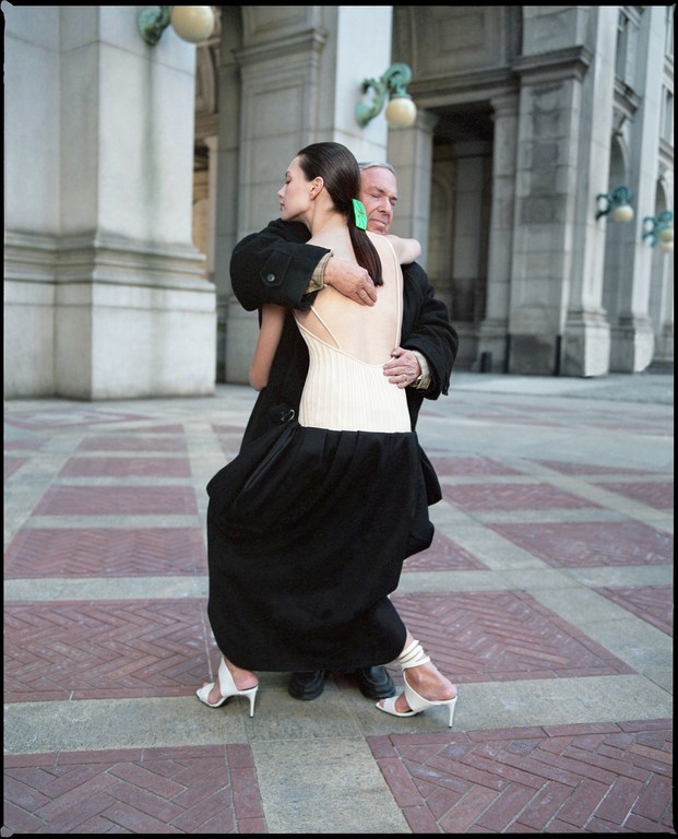 Fashion editorial shot of adult woman in couture dress embracing an older man outdoors against an archtectural background.