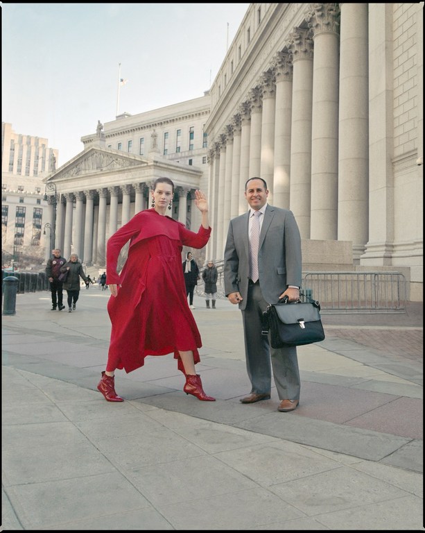 Fashionable woman in a red dress and boots poses next to a suited man with a briefcase in front of a building with columns.