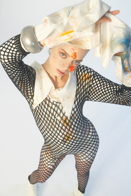 Adult woman in black fishnet bodysuit with white boots, a white collared shirt, and white cloth covered in paint on a white backdrop.