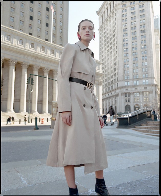 Model in beige trench coat and black belt standing on a paved street surrounded by classical buildings.