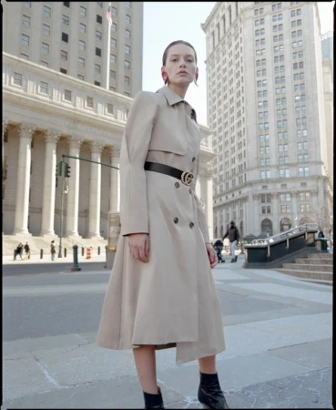 Model in beige trench coat and black belt standing on a paved street surrounded by classical buildings.