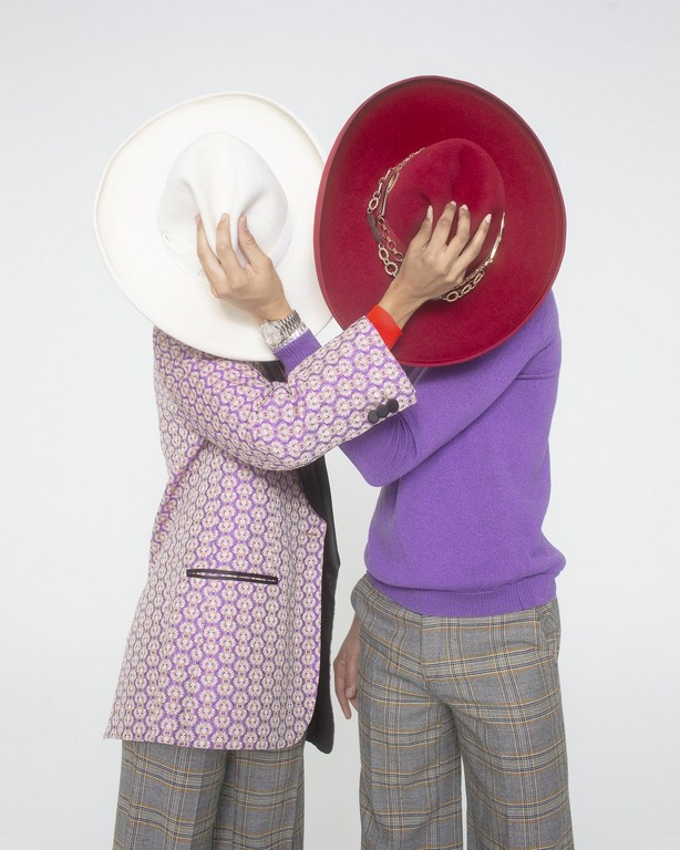 Two people stand closely together, obscuring their faces with a large white and red hat, wearing blazers and patterned pants on a white backdrop.